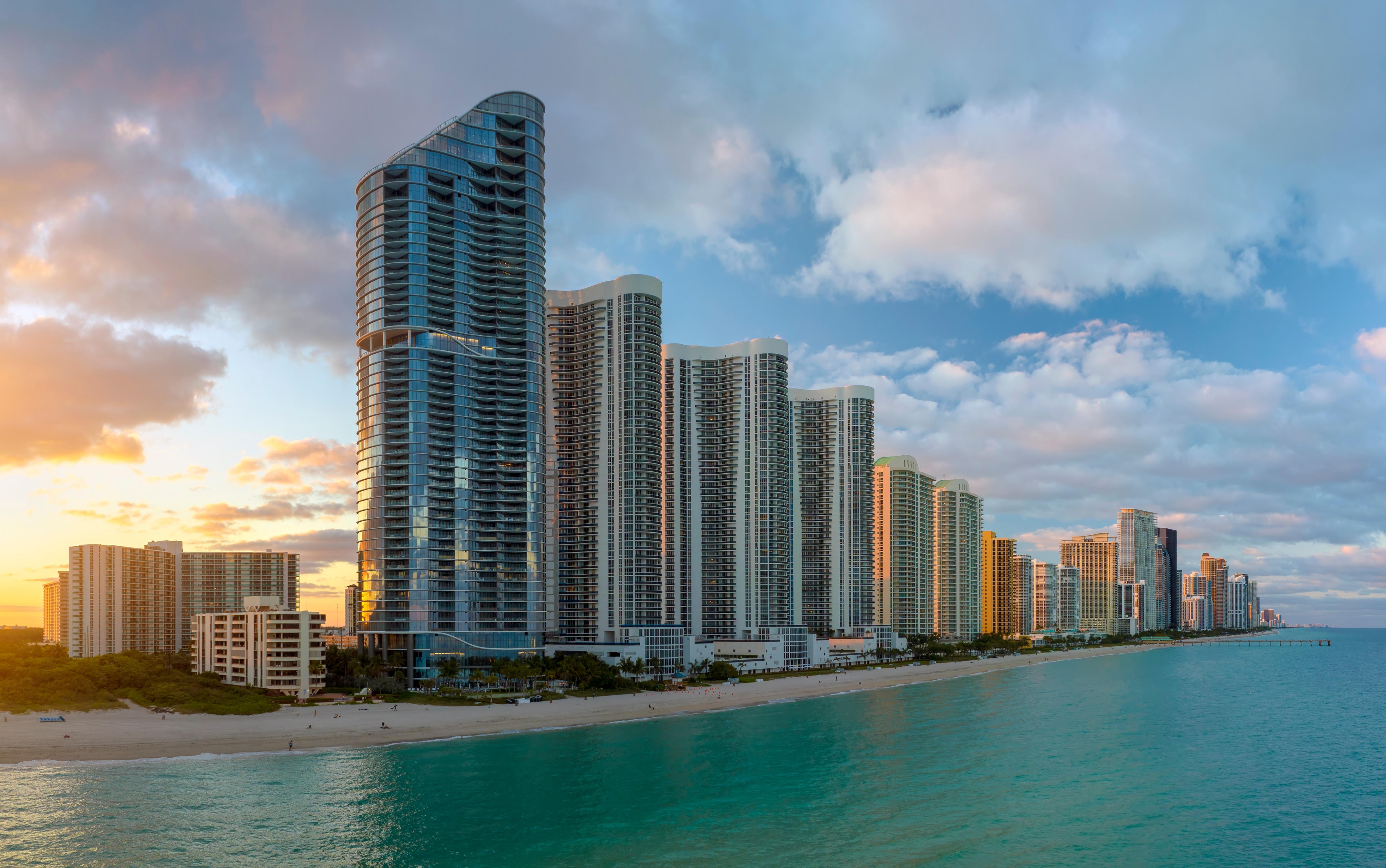 A panoramic view of Sunny Isles Beach, Florida, at sunset, showcasing a stunning row of modern high-rise buildings along the shoreline. The golden sunlight reflects off the glass facades of the skyscrapers, while the turquoise ocean gently laps against the sandy beach. The sky is partly cloudy with soft pastel tones, adding to the serene and luxurious coastal atmosphere.