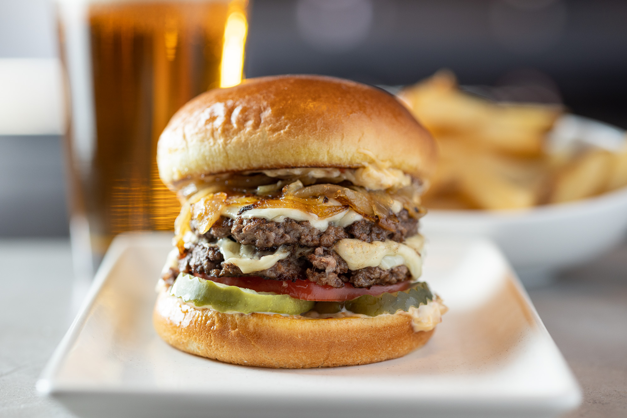 Two patty burger, on a fluffy bun cheese melting on the edge, crispy onions and pickles. On a  plate with a beer and French fries in the background
