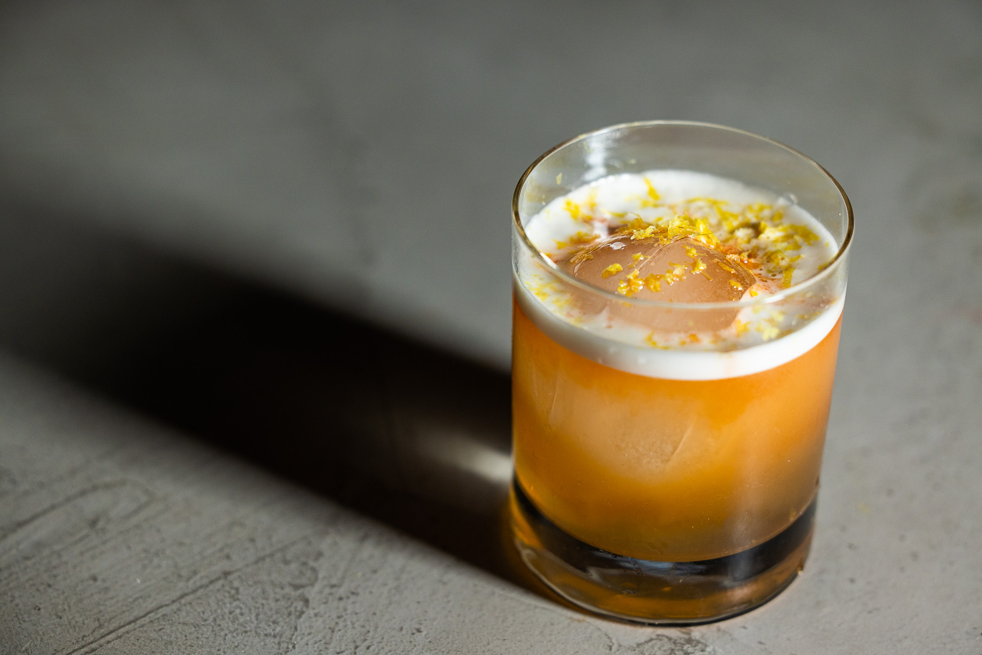 Orange cocktail with foam and lemon zest set on top with a circular ice cube in a short double old fashioned glass 