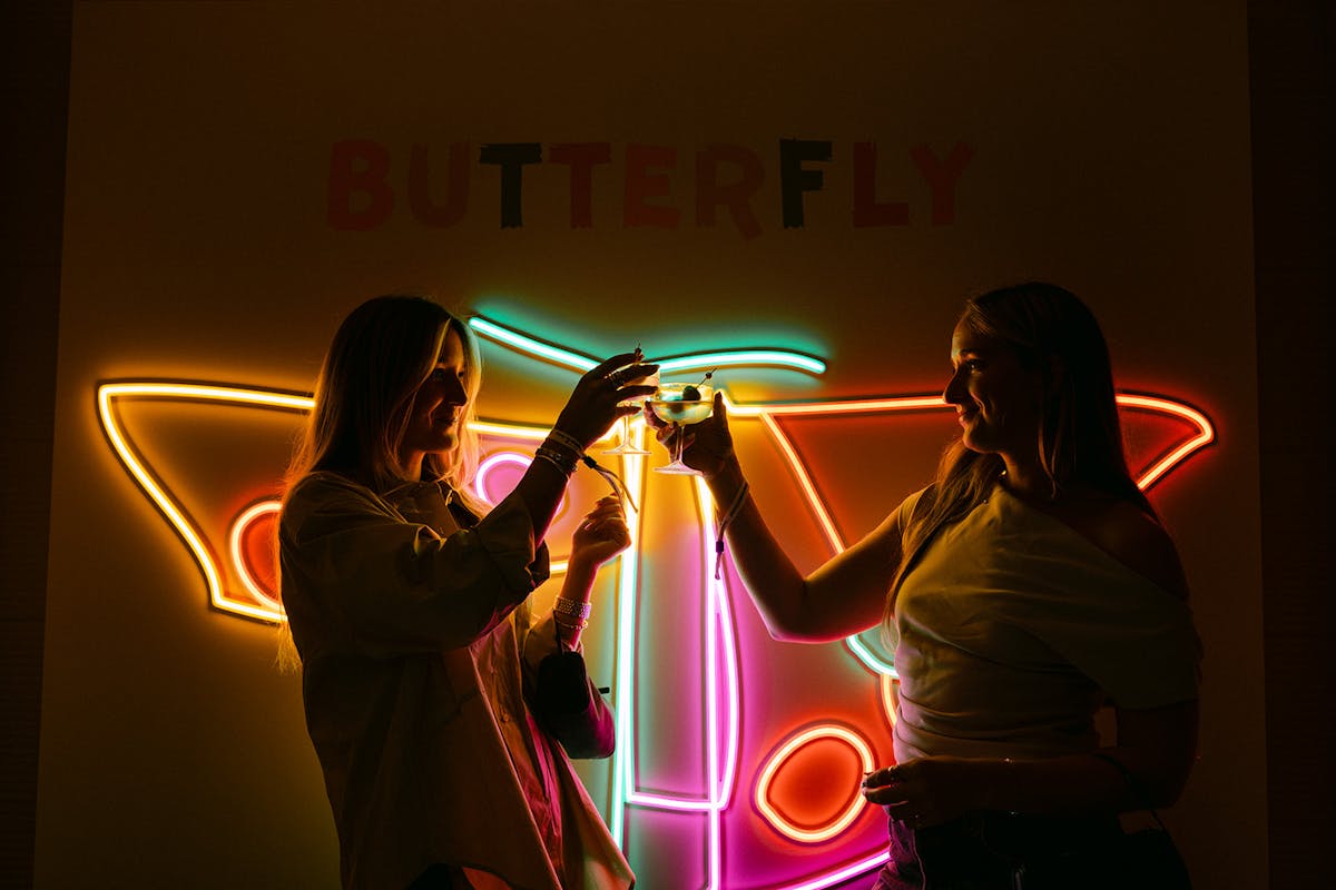 neon butterfly sign and two people holding up cocktails.