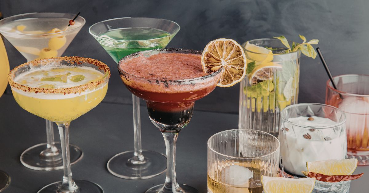 An assortment of colorful spring cocktails, including a yellow margarita with a tajin rim, a red frothy margarita with a dark salt rim and dried lemon slice, a green martini, and a tall mint mojito, all on a dark counter.