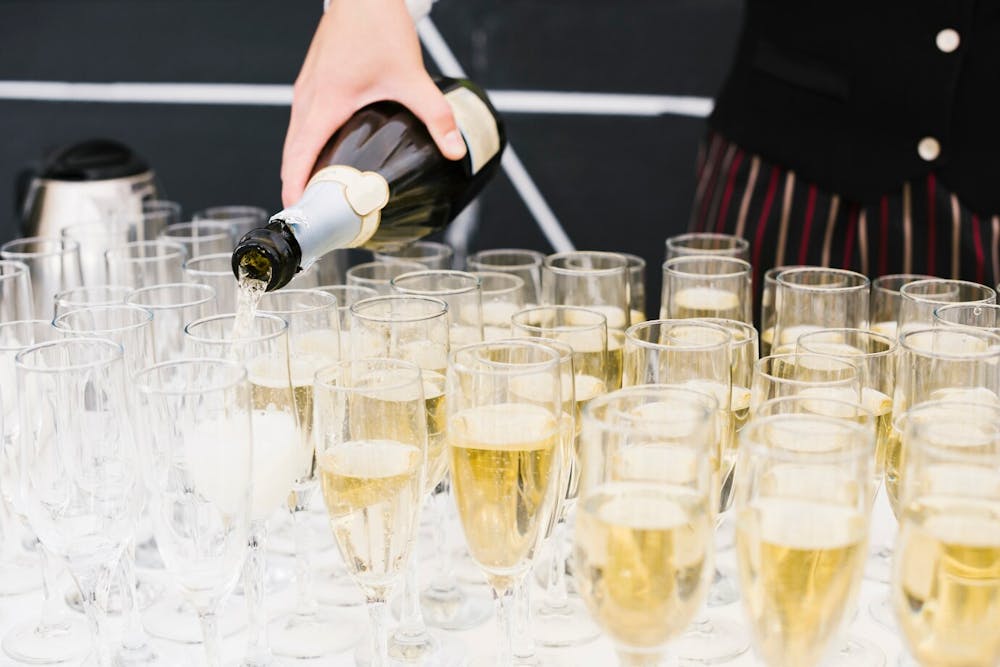 An employee of an alcohol catering company pouring champagne into numerous champagne glasses sitting on a table with white table cloth.