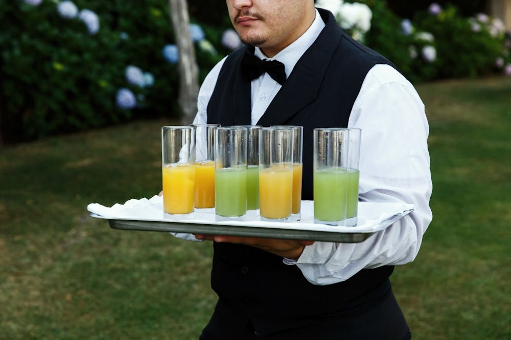 An employee of an alcohol catering company carrying eight tall glasses of yellow and green cocktails on a silver tray lined with white paper towels.