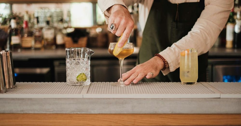 A bartender in a green apron carefully expresses a lemon peel over a golden-colored cocktail in a coupe glass at a modern bar.