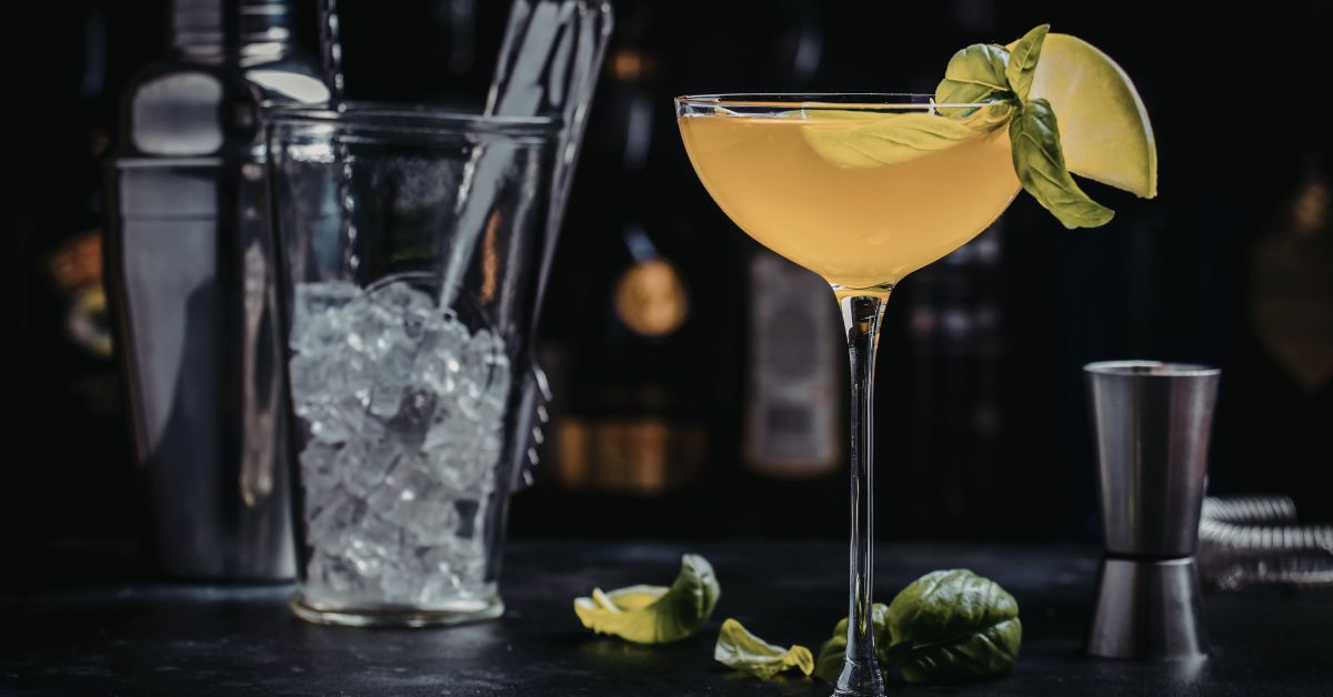 A close-up of a bright yellow spring cocktail in a coupe glass, garnished with fresh basil leaves and a lime wedge, with bar tools in the background.