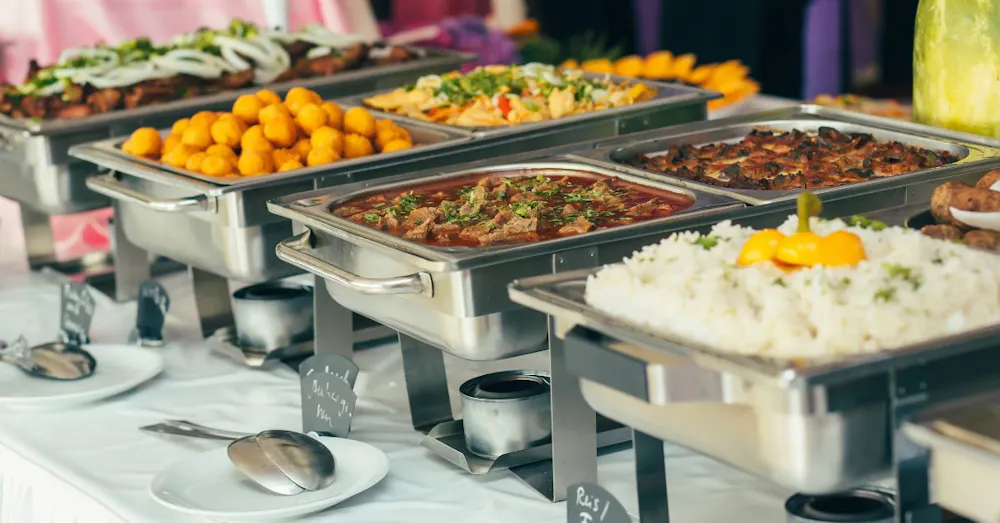 A professional wedding buffet line featuring stainless steel chafing dishes filled with rice, meat entrees, and vegetable sides on a white tablecloth.