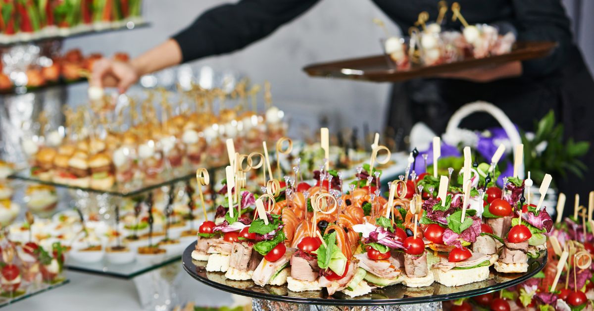A close-up of a catering display featuring gourmet hors d'oeuvres with salmon, cherry tomatoes, and microgreens on small bread bases with decorative picks.