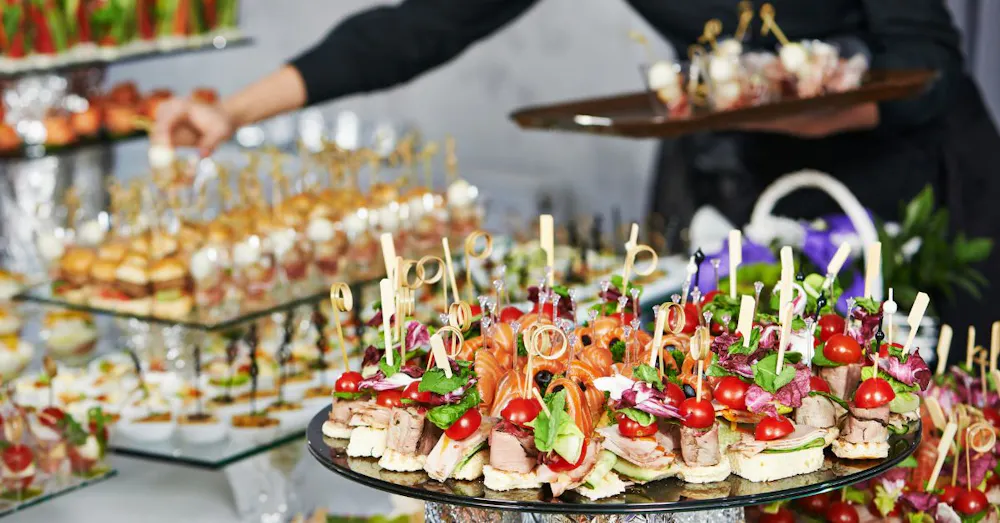 A close-up of a catering display featuring gourmet hors d'oeuvres with salmon, cherry tomatoes, and microgreens on small bread bases with decorative picks.