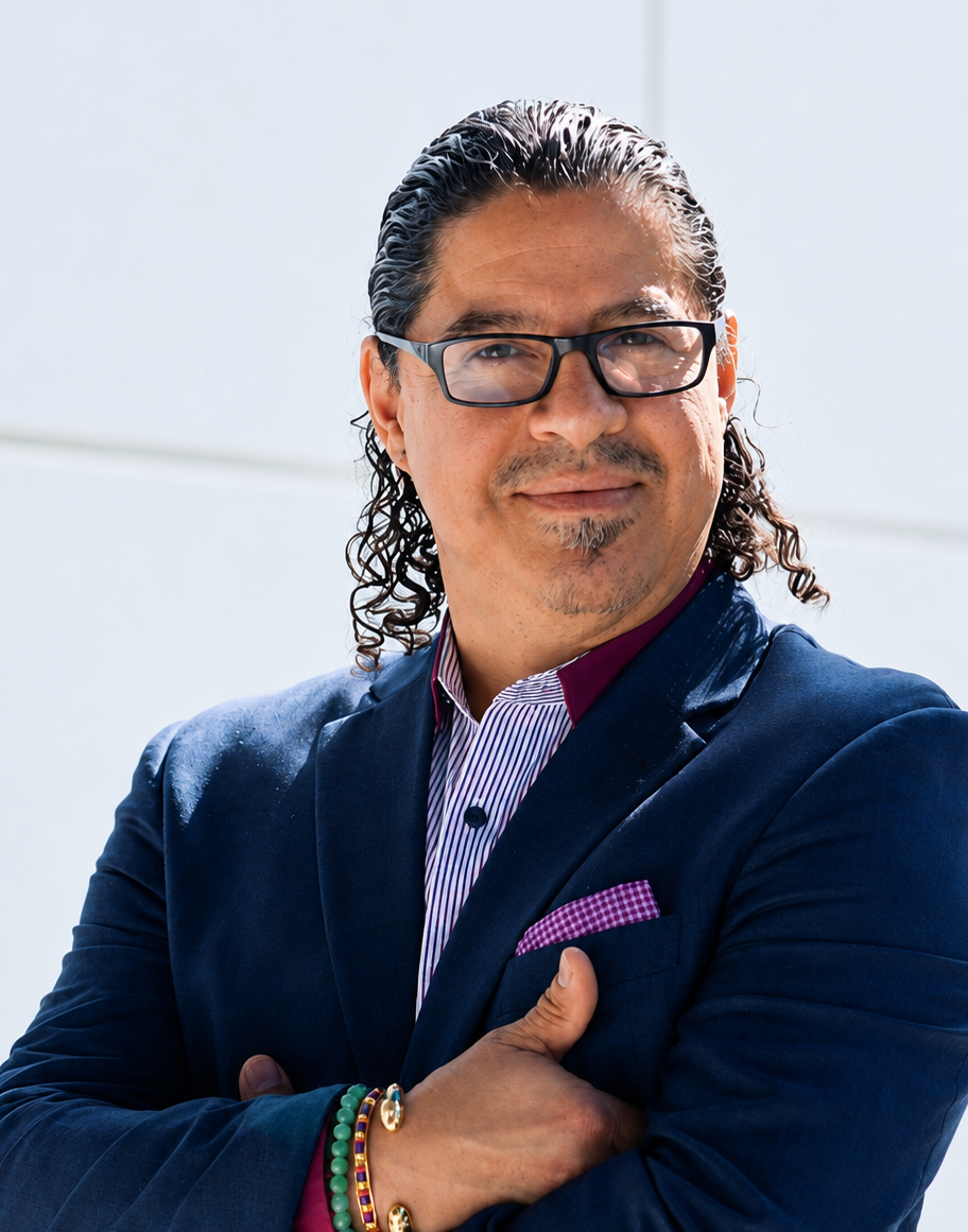 Professional headshot of a man wearing a navy blazer, striped dress shirt and glasses, standing with his arms crossed against a bright white background.