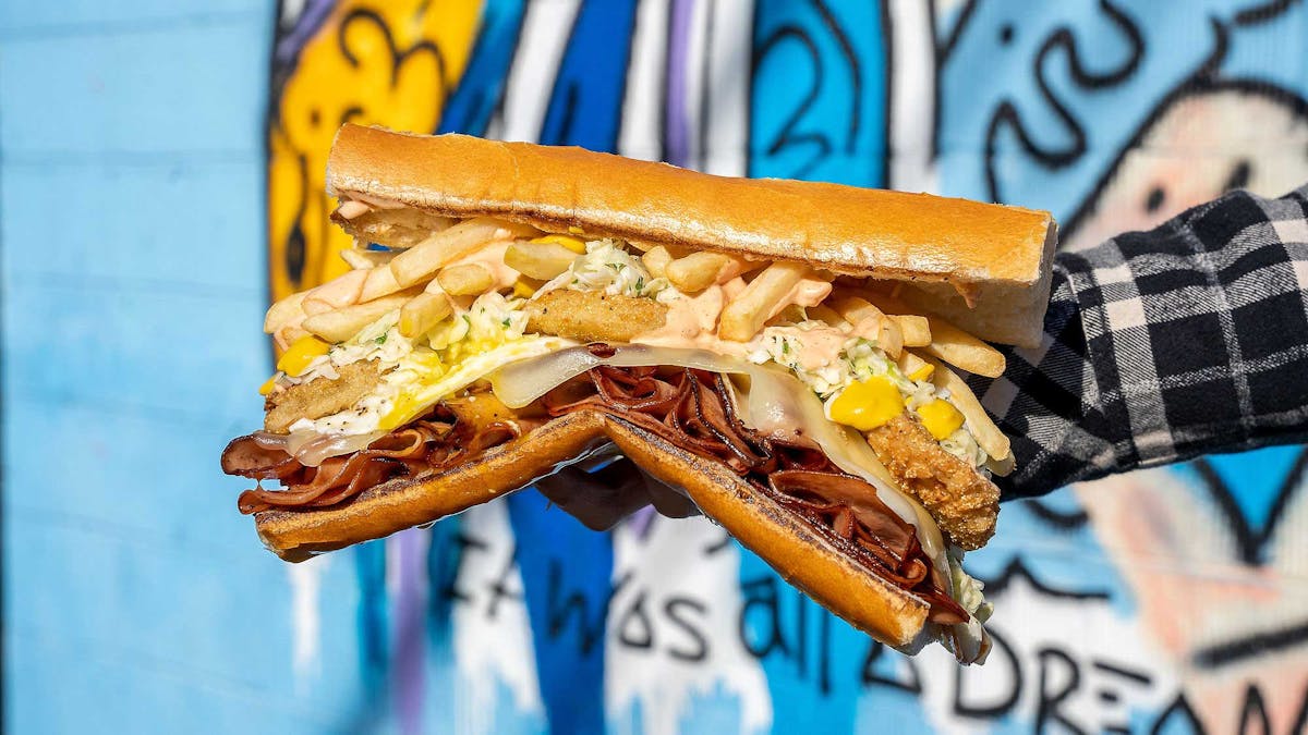 Fat Sal’s Deep South sandwich with roast beef, fried chicken, fries, coleslaw, cheese, and special sauce on a hero roll, held in front of street art
