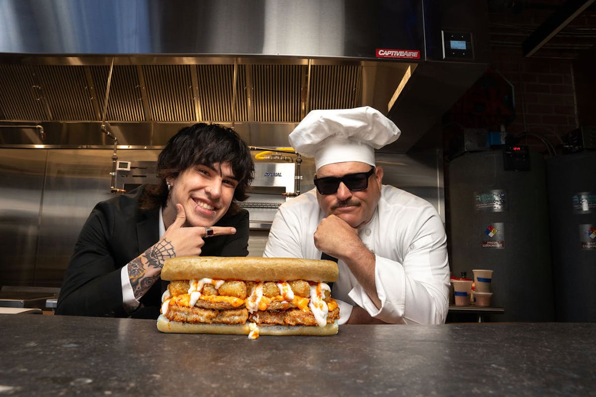 Jake Weber and chef Sal posing with an overstuffed sub sandwich in a commercial kitchen, showcasing bold flavors and creative fast food culture.