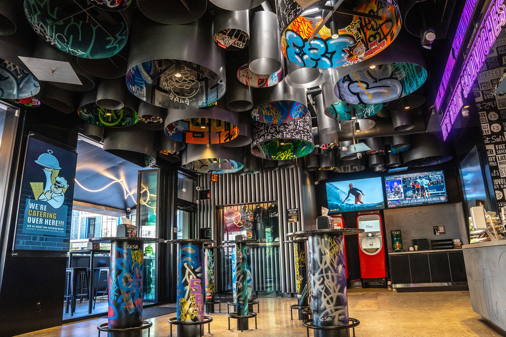 Interior of Fat Sal’s Culver City, featuring graffiti-covered tabletops, vibrant ceiling art made from cylindrical installations, neon signage, and flat screens above the counter.