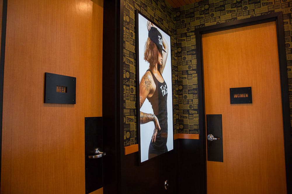 Inside Fat Sal’s Encino, showing the men’s and women’s restrooms with wood doors, black signage, and a bold wall graphic of a tattooed woman in Fat Sal’s gear.