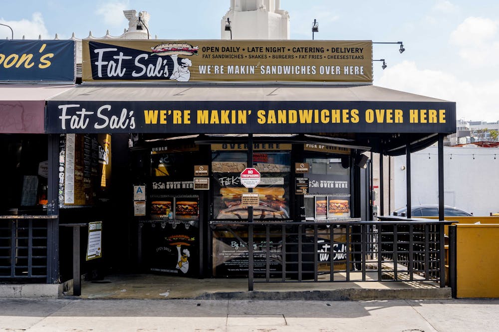 The front entrance of Fat Sal’s Westwood with bold signage reading “We’re Makin’ Sandwiches Over Here,” menu posters in the windows, and a black railing around the patio area.