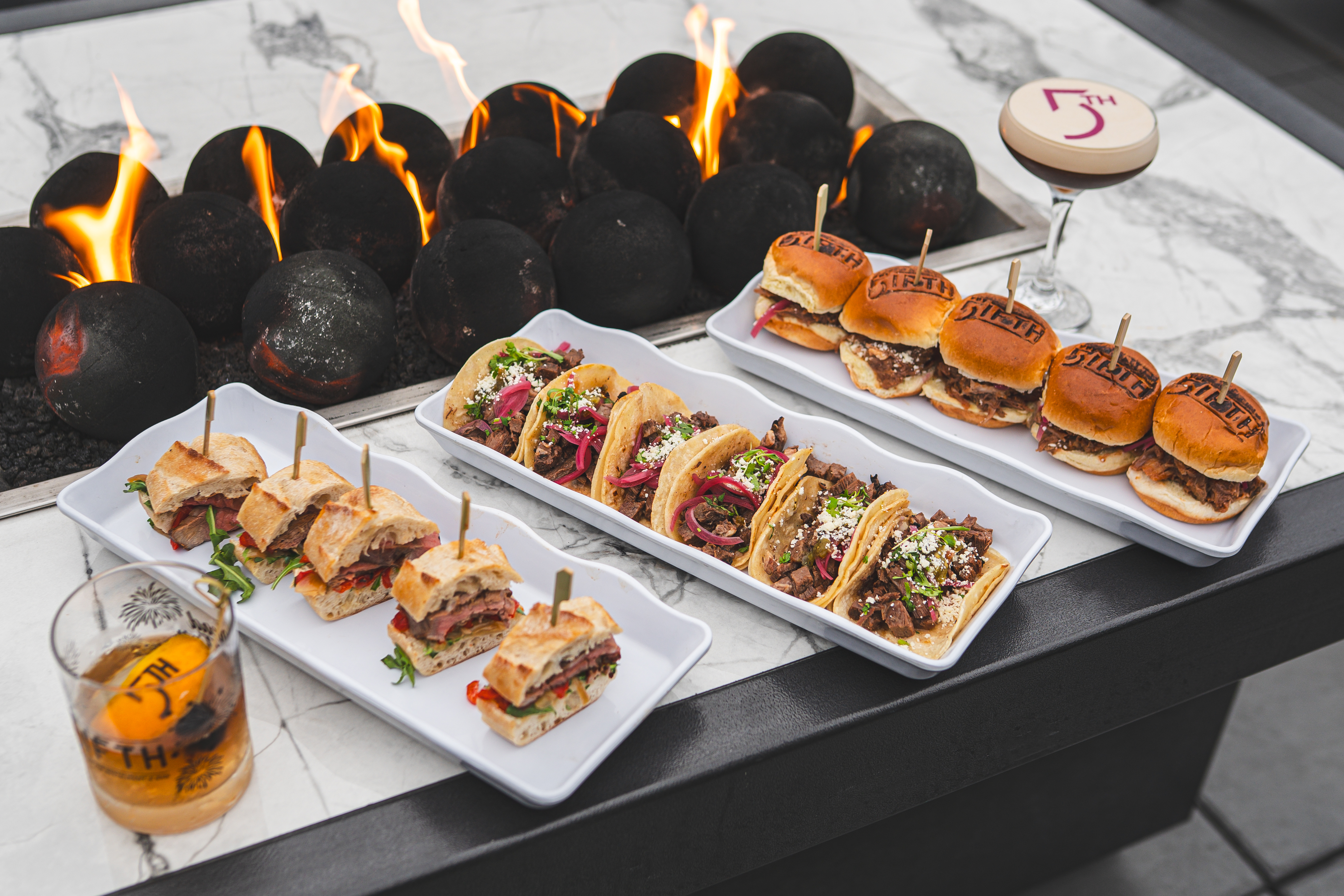 Plates of mini sliders, tacos, and sandwiches stationed on a fireplace table next to two cocktail drinks