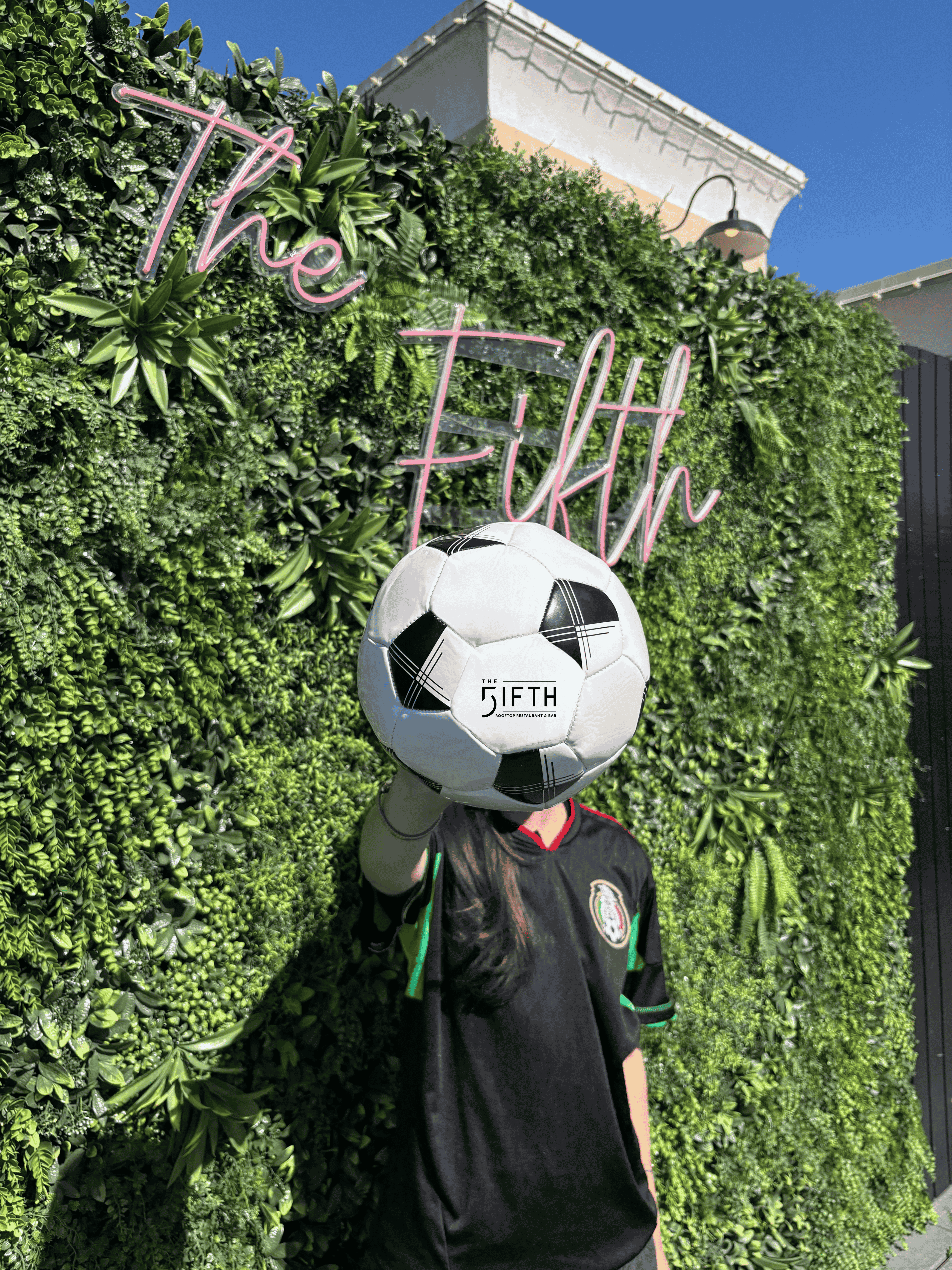 **Alt text:** A fan wearing a Mexico soccer jersey holds up a soccer ball in front of a green hedge backdrop featuring a neon sign.