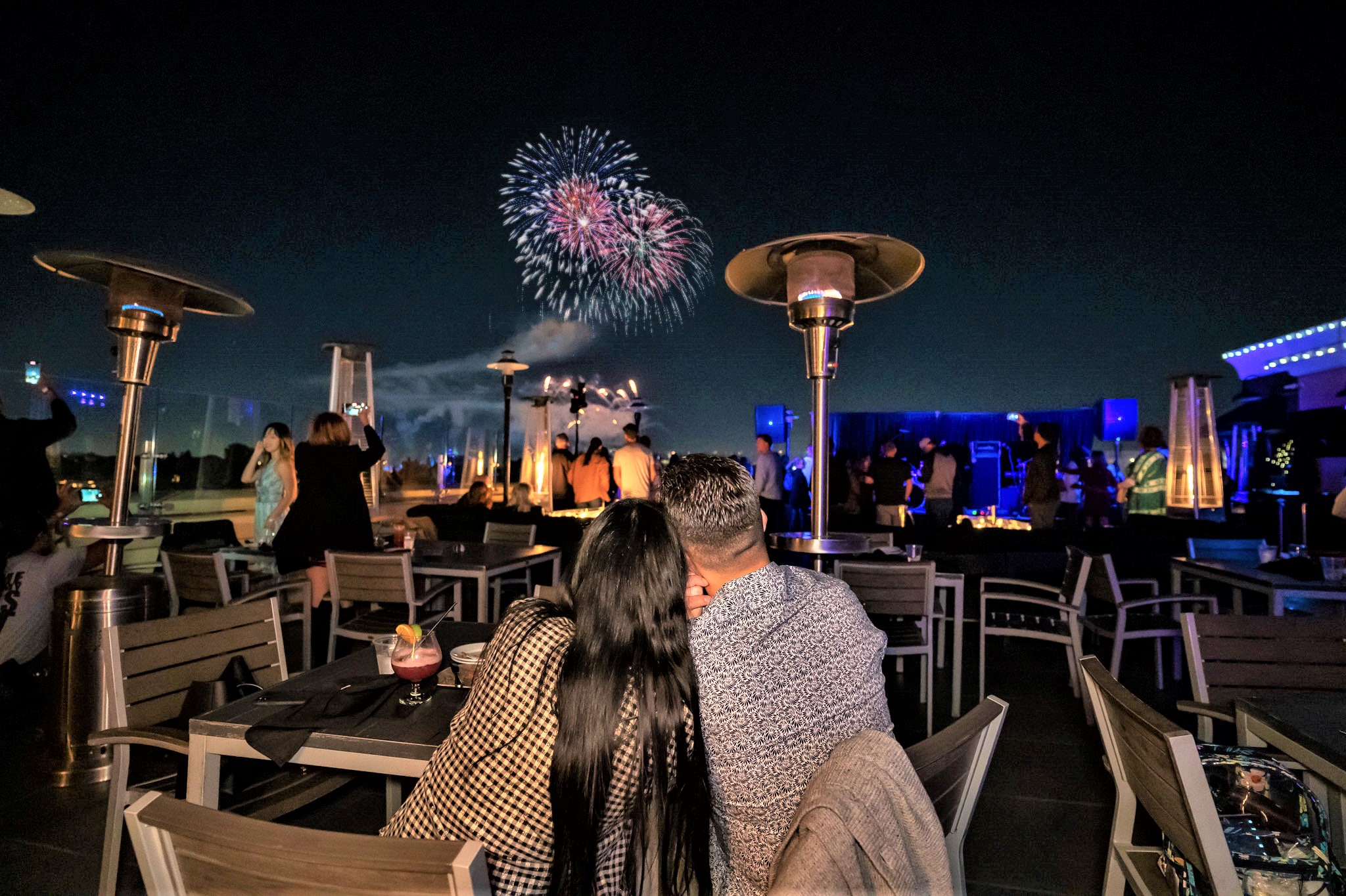 A couple sitting at a rooftop restaurant table at night watching purple and pink 4th of July fireworks over Anaheim, with outdoor heaters and a lively crowd at The FIFTH Rooftop Bar.
