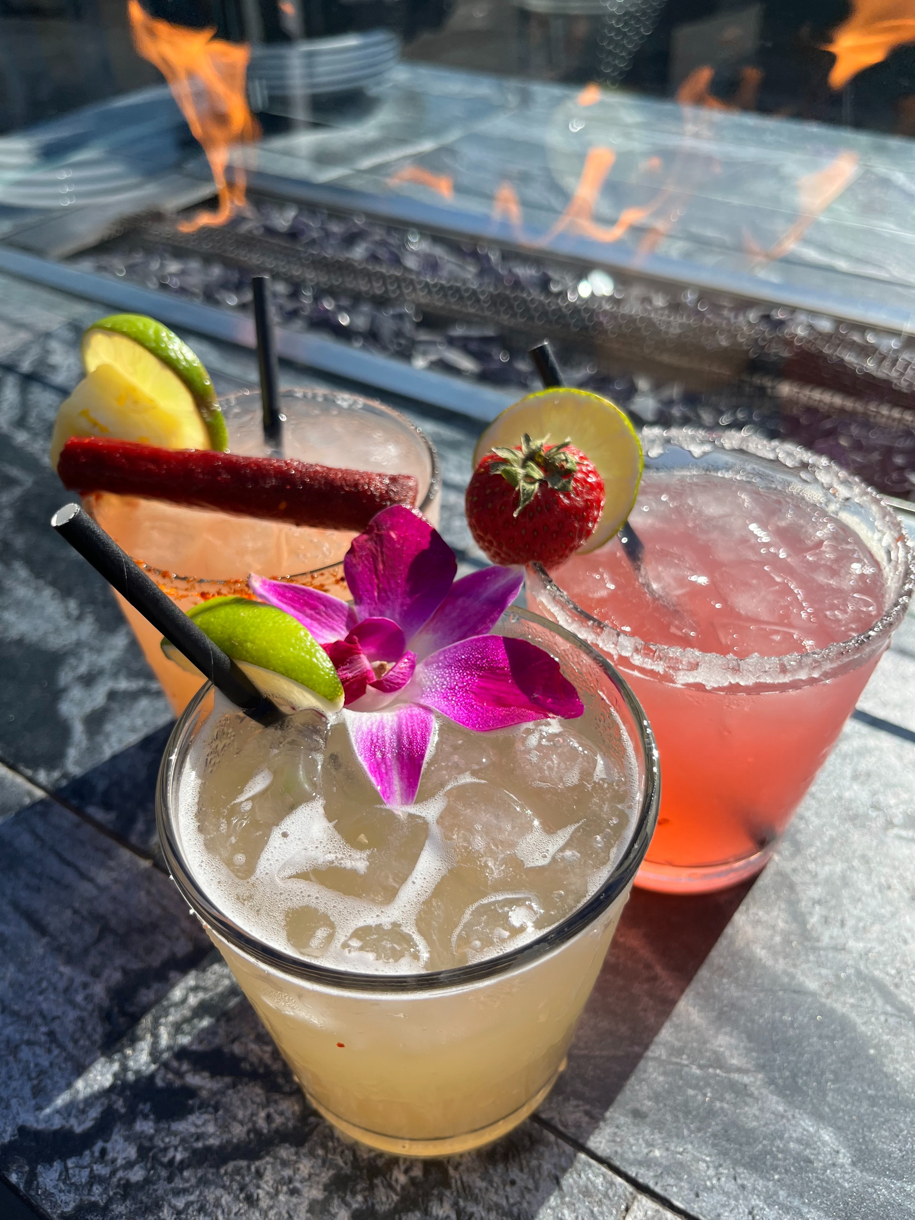 Craft margaritas with fresh fruit garnishes and a tropical flower served beside a rooftop fire pit at The FIFTH rooftop bar in Anaheim.