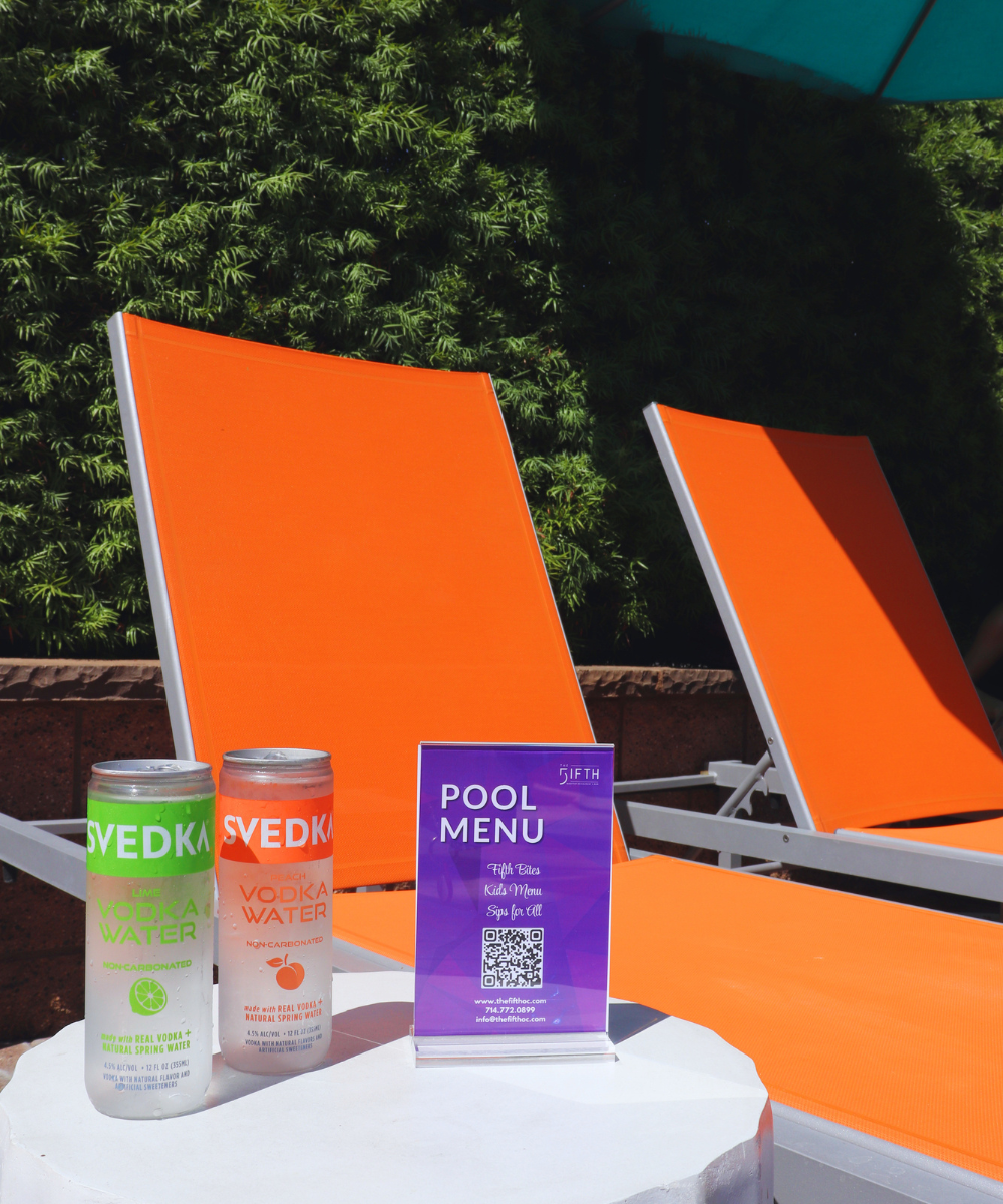 Two canned cocktails sitting on a white countertop next to an orange lounge chair with The FIFTH's pool bar menu.