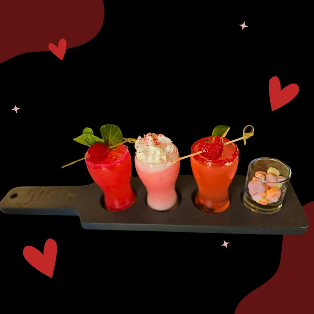 A cocktail flight featuring a red cocktail with cherry garnish, a pink cocktail with whipped cream and sprinkles, a red cocktail with red sugar rim and red candy garnish, and a glass of heart candies