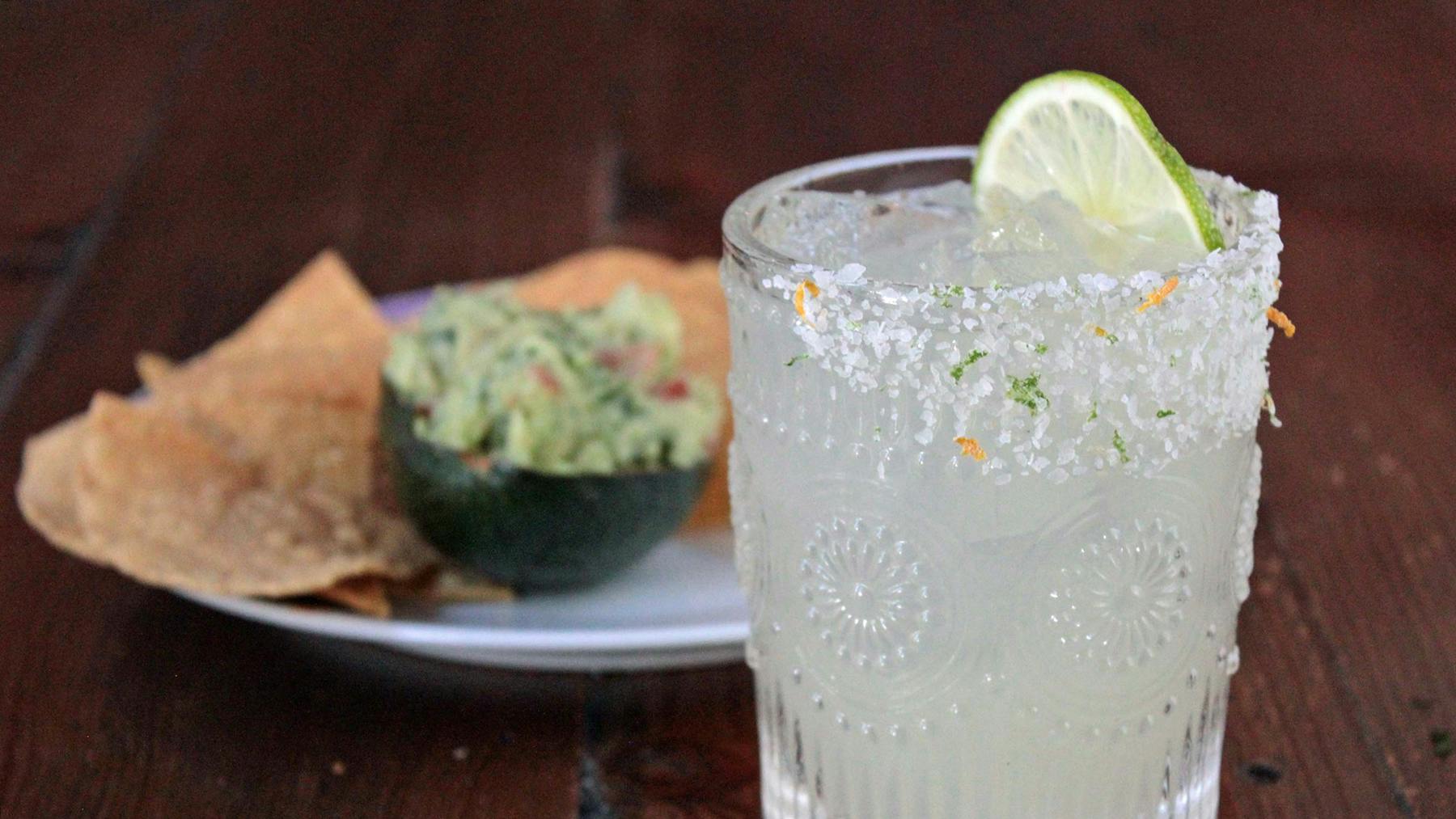 a glass of margarita with lime and guacamole in an avocado with chips on a plate in the back