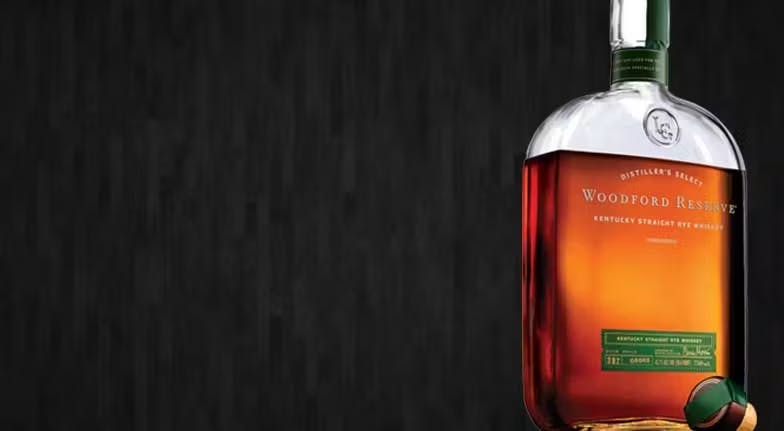 A bottle of Woodford Reserve Kentucky Straight Rye Whiskey against a dark textured background. The bottle is half-filled with amber liquid, conveying elegance.
