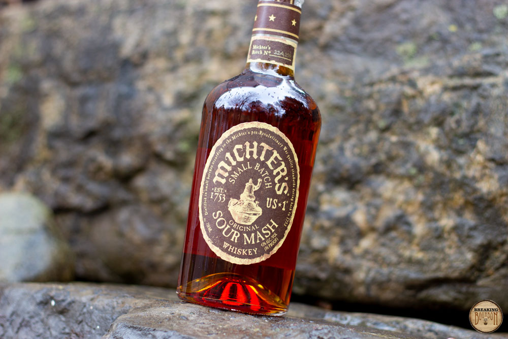 A bottle of Michter's Sour Mash Whiskey is tilted against a rough, stone background. The amber liquid is visible, and the label is prominent.