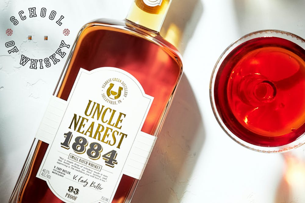 Overhead view of Uncle Nearest 1884 whiskey bottle angled beside a full red cocktail on a light surface. "School of Whiskey" text is seen at the top left.