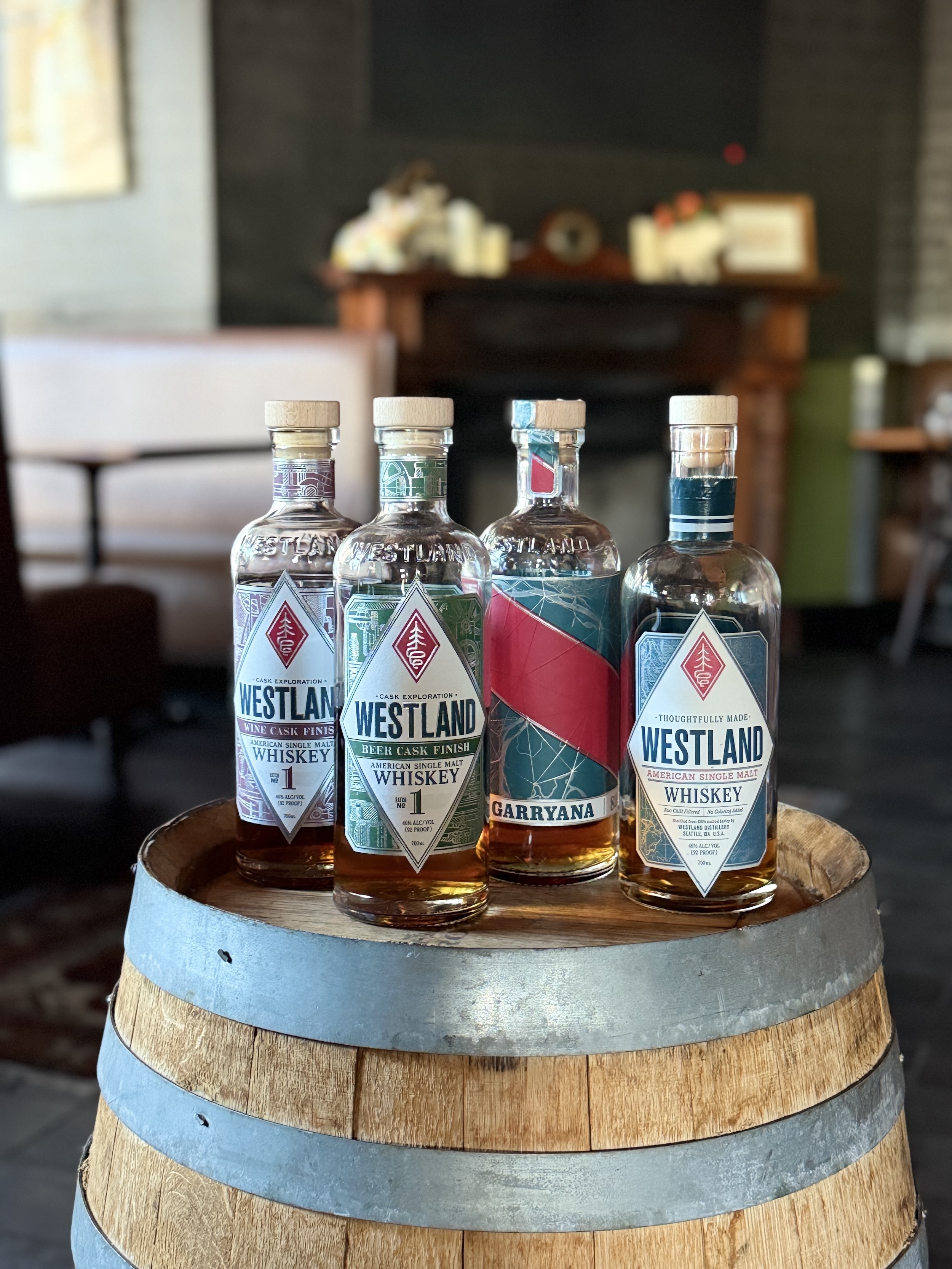 Four bottles of Westland whiskey sit on a wooden barrel in a cozy room. Labels are colorful and distinct, evoking an elegant, inviting atmosphere.