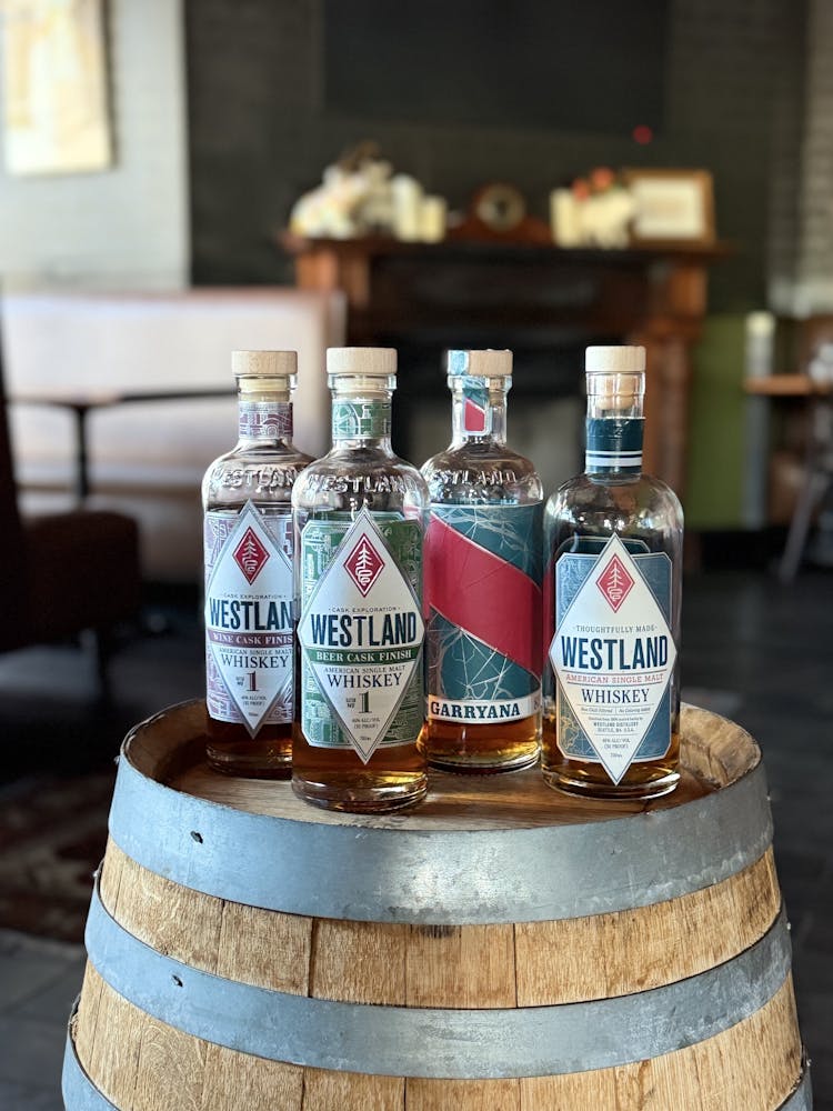 Four bottles of Westland whiskey sit on a wooden barrel in a cozy room. Labels are colorful and distinct, evoking an elegant, inviting atmosphere.