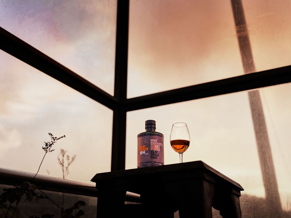 A bottle and glass of whisky sit on a table by a window with a moody sunset sky. The bottle is labeled prominently, adding a rustic ambiance.
