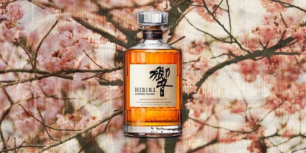 A bottle of Hibiki Japanese Harmony whiskey with an amber hue stands in front of a blurred background of pink cherry blossoms, evoking elegance and tradition.