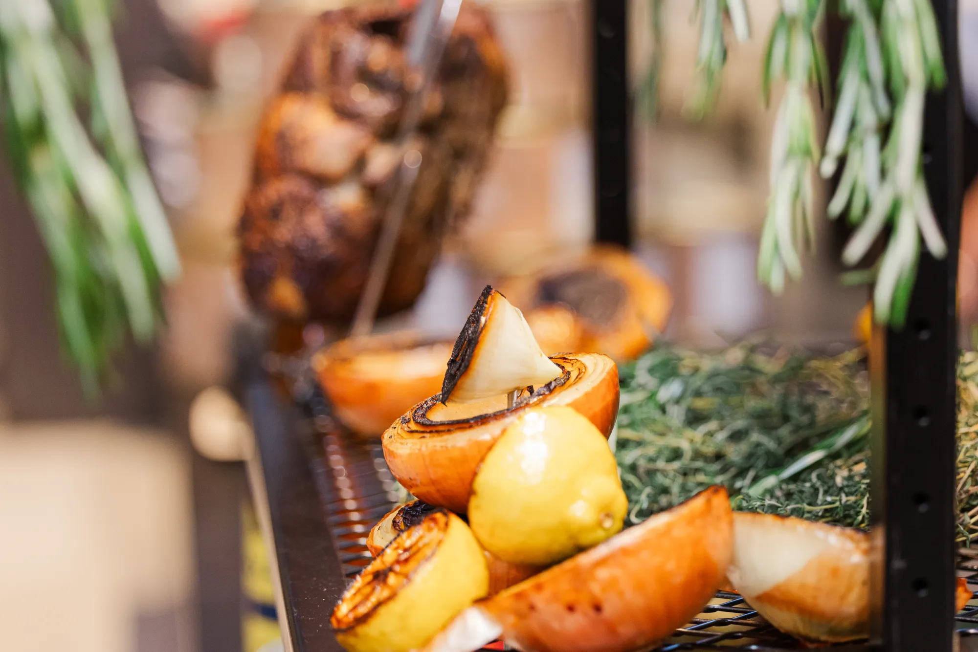 Charred onions and lemons on a grill with hanging herbs and roasted meat in a warm Boston summer setting.