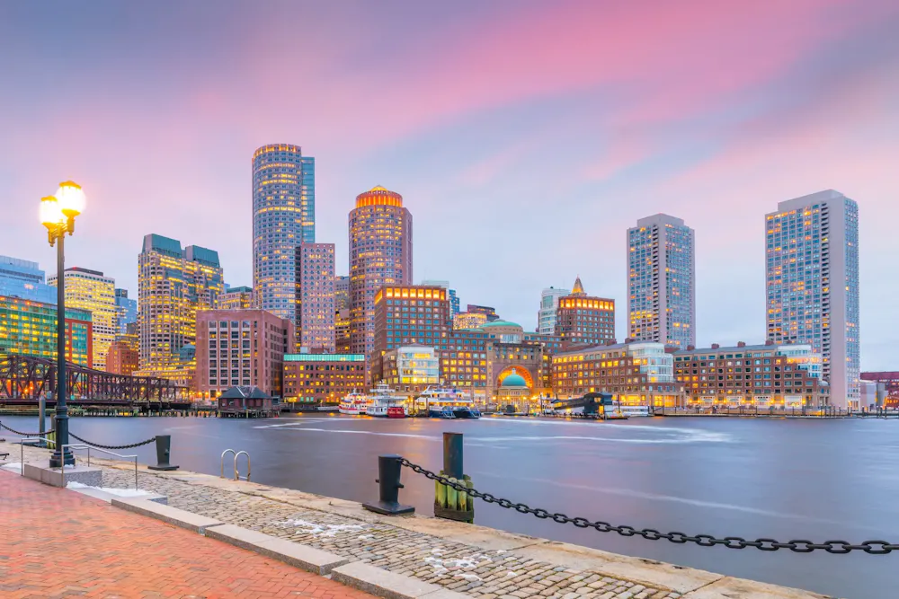 Boston Harbor at sunset with waterfront, bridge, and illuminated skyscrapers in Boston’s Financial District.