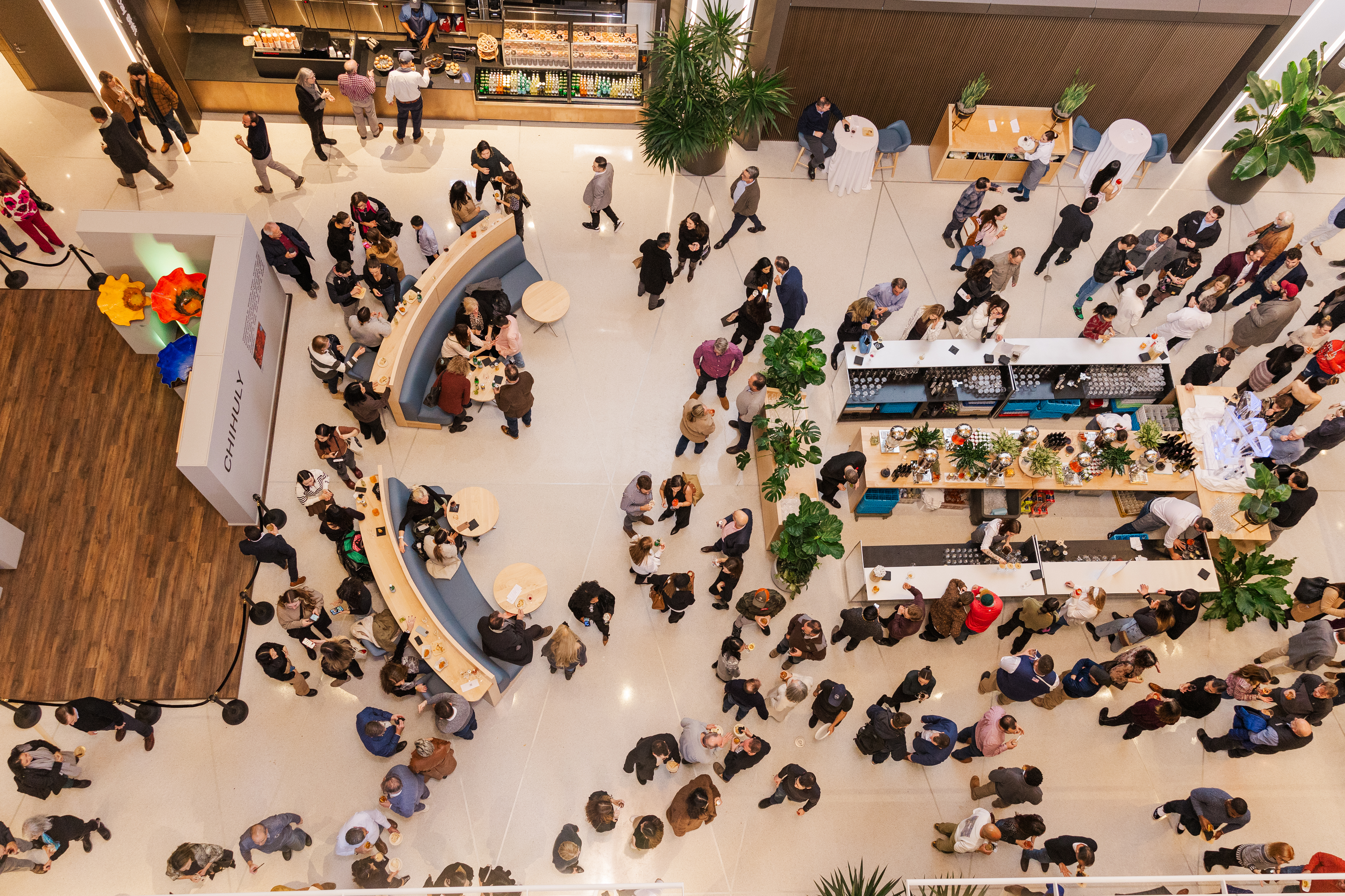 Overhead view of a busy indoor event space where dozens of people are mingling, walking, and standing in line at long buffet tables and a curved seating area.