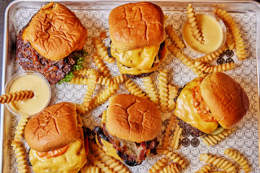 Tray of smashburgers from Big Grin with crinkle cut fries and dipping sauce cups arranged on a patterned liner.