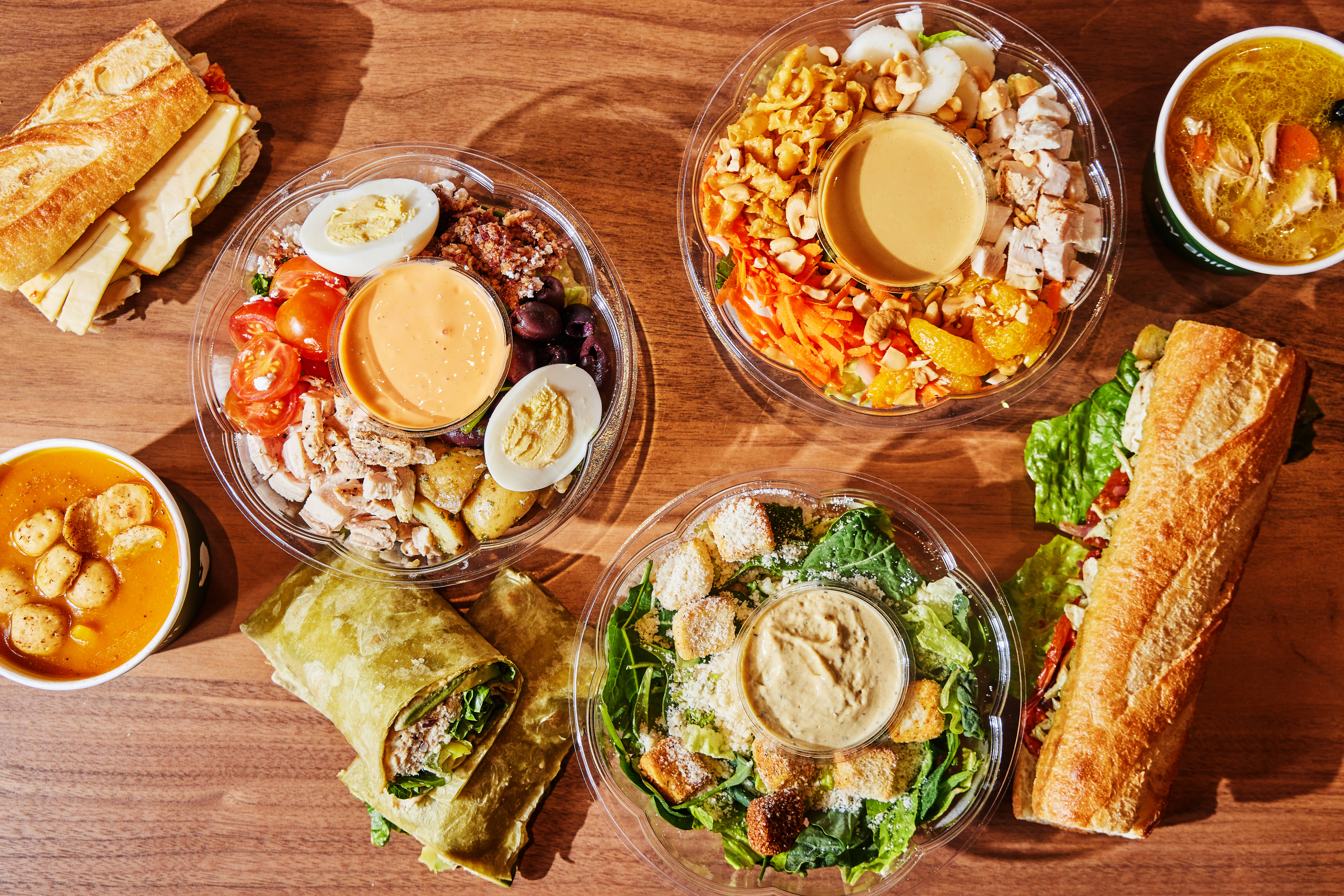 Assorted meals from Day Shift on a wooden table including salads, sandwiches, wraps, and bowls of soup.