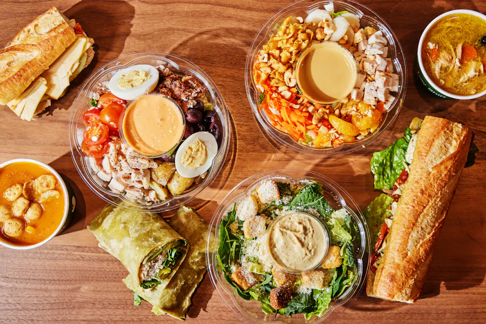 Assorted meals from Day Shift on a wooden table including salads, sandwiches, wraps, and bowls of soup.