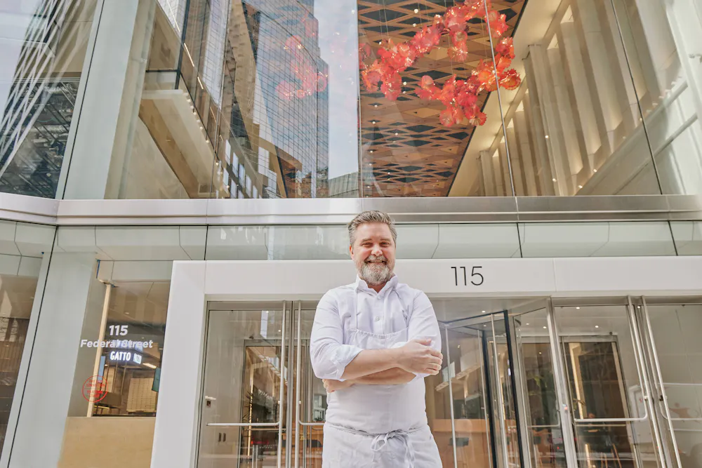 Chef John Fraser stands outside the modern glass building housing The Lineup, smiling with arms crossed.