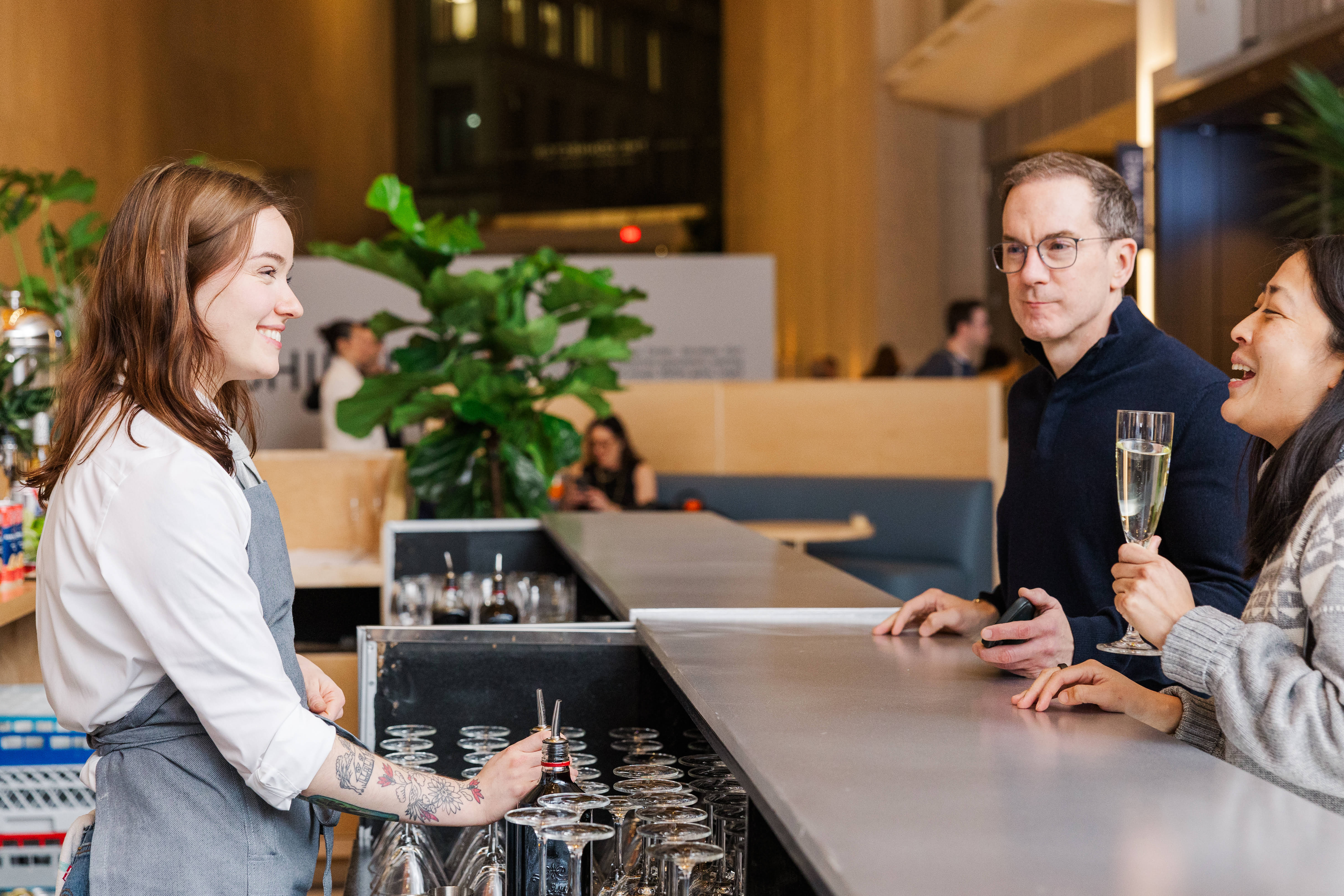 Bartender smiling while serving two guests at a modern bar as they chat and hold a glass of sparkling wine.