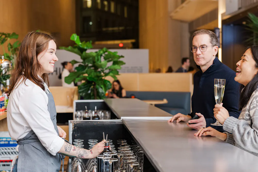Bartender smiling while serving two guests at a modern bar as they chat and hold a glass of sparkling wine.