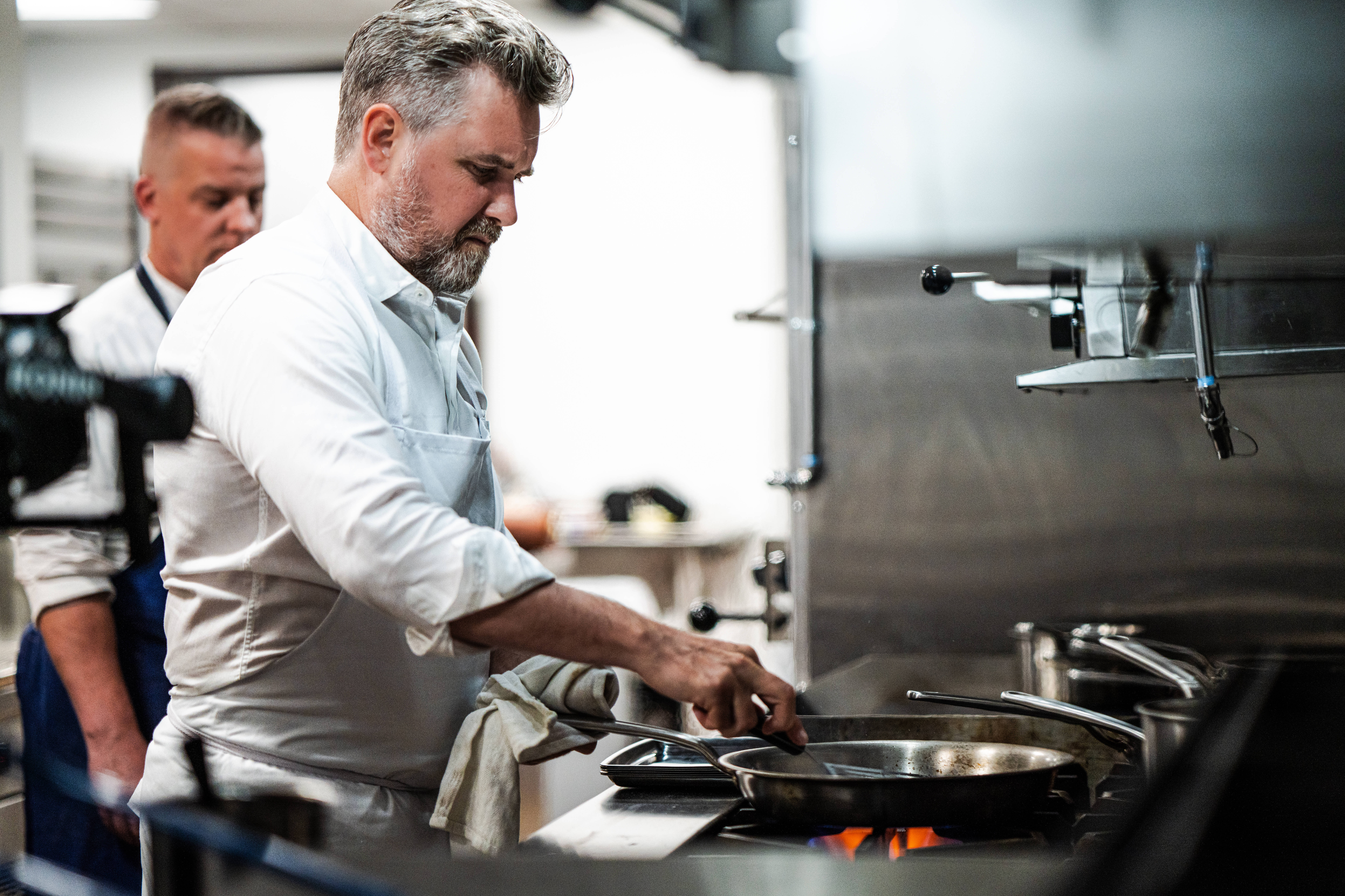 Chef John Fraser cooks at a stovetop in his food hall kitchen while another chef works in the background.