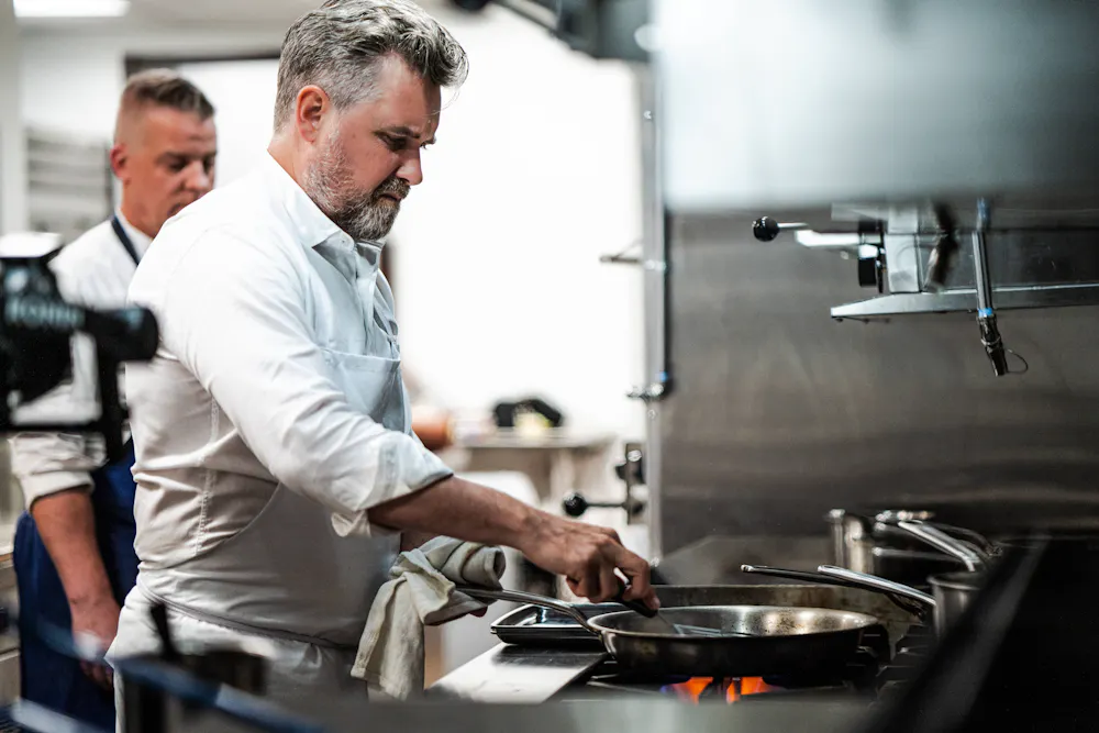 Chef John Fraser cooks at a stovetop in his food hall kitchen while another chef works in the background.