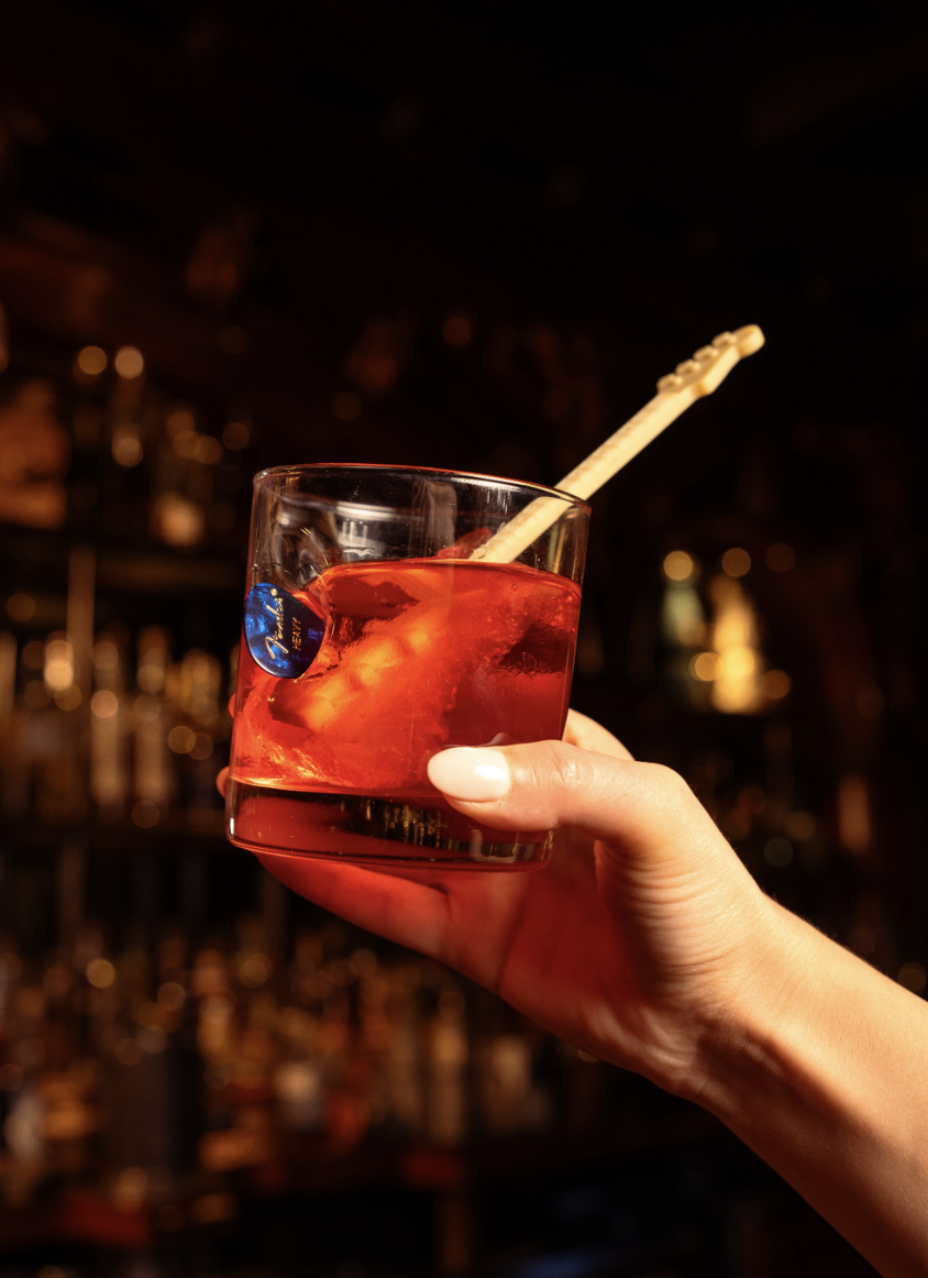 A hand holding a red cocktail over ice, garnished with a guitar-shaped stirrer, at The Barbershop Las Vegas speakeasy
