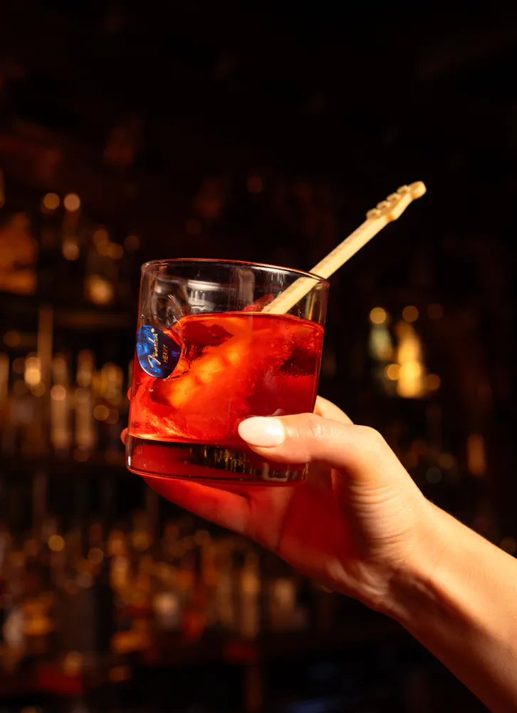 A hand holding a red cocktail over ice, garnished with a guitar-shaped stirrer, at The Barbershop Las Vegas speakeasy