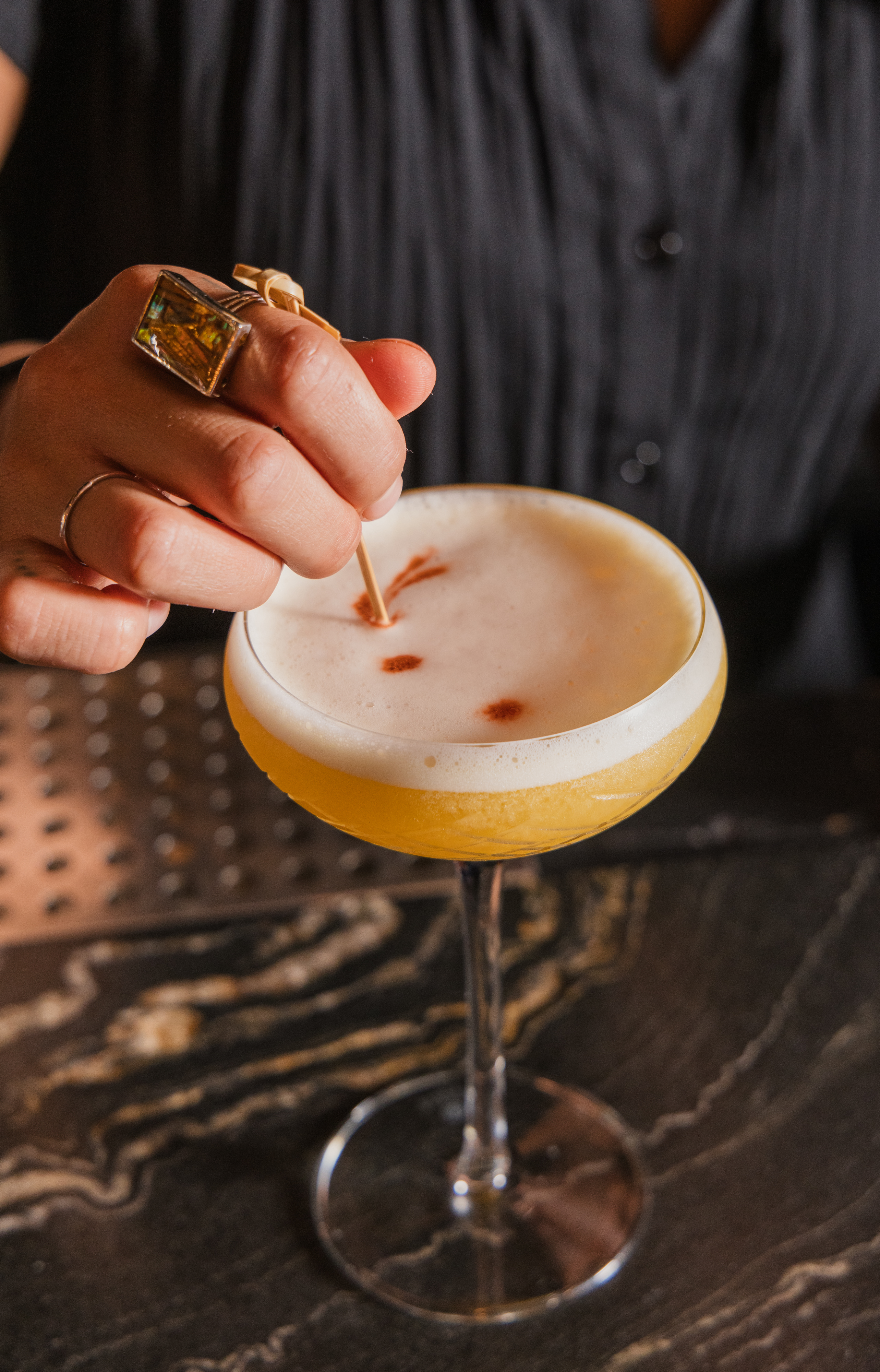 A bartender's hand adding a finishing garnish to a craft cocktail with a frothy top at KAMA