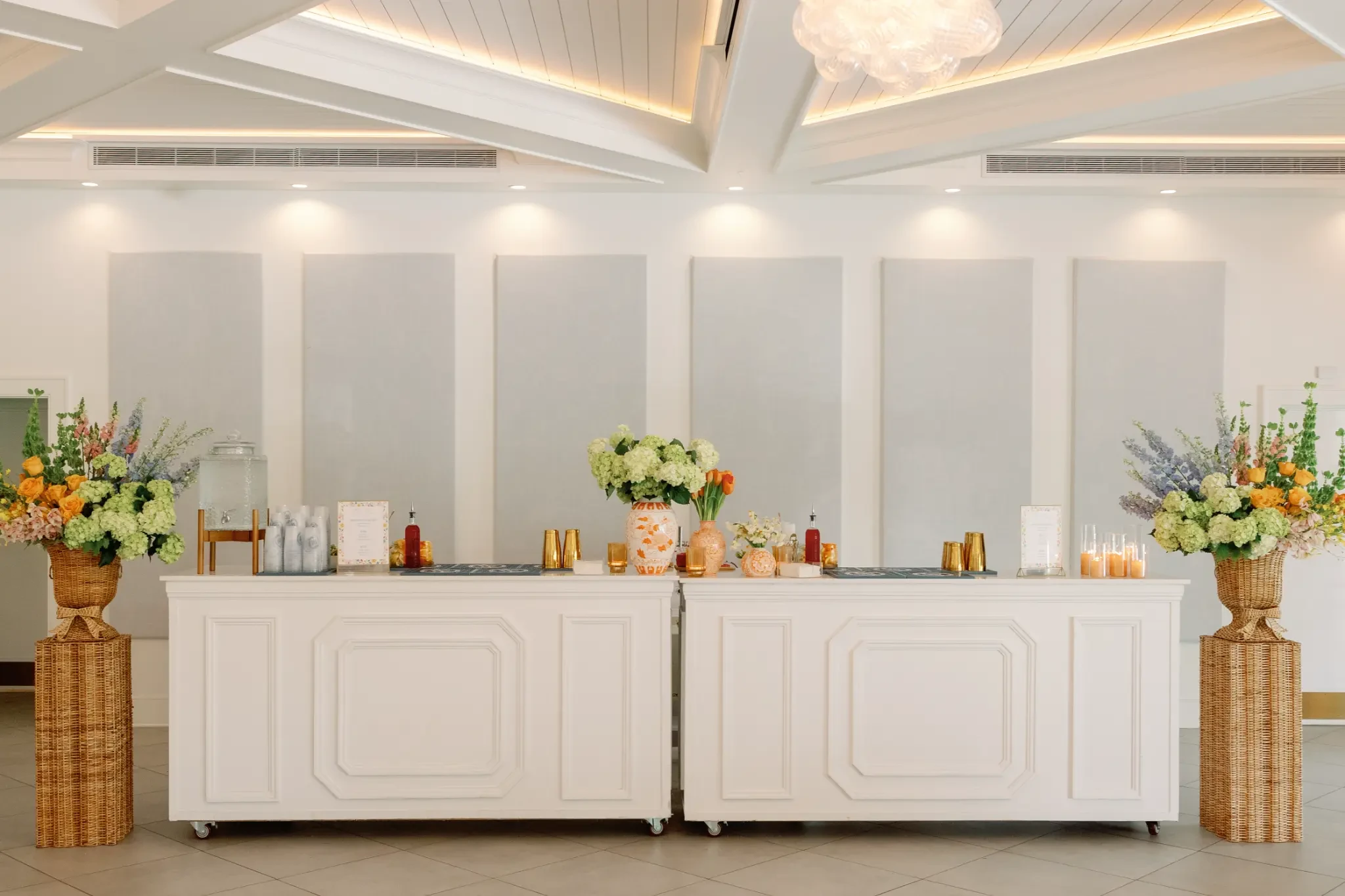 Two white indoor bars with colorful flowers, glassware and gold cocktail shakers beneath The Waverly's vaulted ceiling