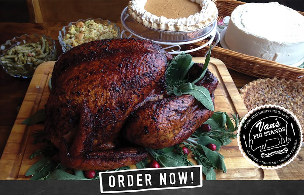Roasted turkey on a wooden cutting board garnished with herbs and cranberries, surrounded by Thanksgiving sides, including green bean casserole, stuffing, pies, and a cake. Van's Pig Stands logo is in the bottom right corner. Along with the words ORDER NOW! bottom center.