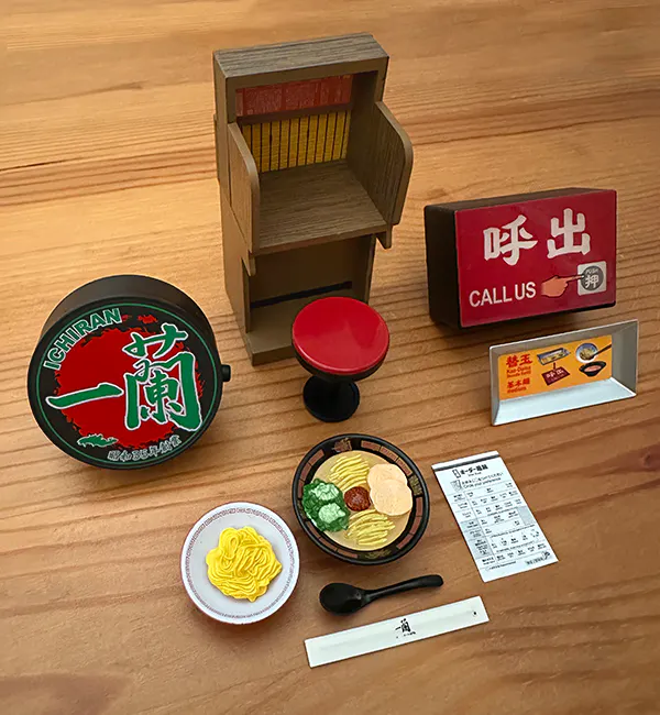 Collection of the ICHIRAN miniature capsule toys: ICHIRAN light up logo magnet, Ramen Focus booth and stool, Call button and Kae-Dama plate, Classic Tonkotsu Ramen with a bowl of Kae-Daman and order form, renge ramen spoon, and chopsticks in paper sleeve