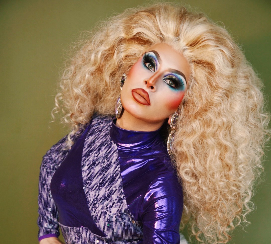 A drag performer with voluminous blonde curls poses dramatically against an olive-green backdrop. They wear a shimmering purple sequin bodysuit, white thigh-high boots, and large rhinestone jewelry with bold, colorful stage makeup.